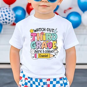 May include: White short-sleeved t-shirt with the text "Watch Out Third Grade here I come!" in colorful bubble letters. The shirt has a pencil graphic with the name "Daniel" on it. The child is wearing blue sunglasses and checkered shorts.