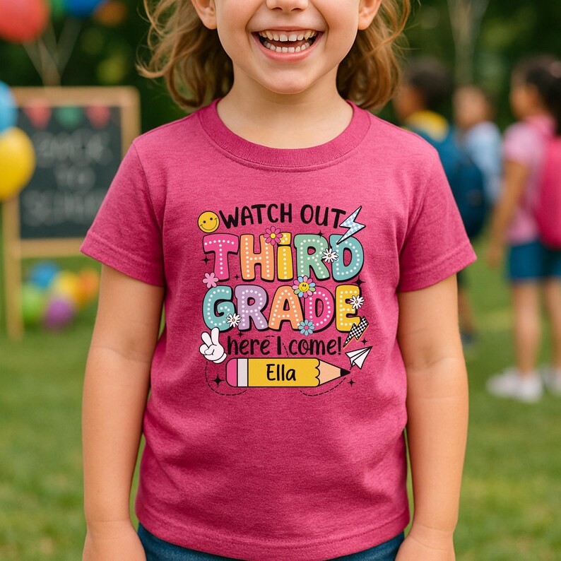 May include: A magenta short-sleeved t-shirt with the text "Watch Out Third Grade Here I Come! Ella" in colorful bubble letters and graphics. The shirt features a smiling face, flowers, a peace sign, a pencil, and a paper airplane.