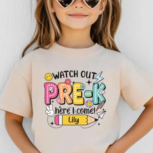 May include: A beige t-shirt with the text "WATCH OUT PRE-K here I come!" in colorful letters and graphics. The name "Lily" is printed on a yellow pencil graphic. The shirt is designed for preschool.