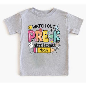 May include: A gray short-sleeved t-shirt with the words "WATCH OUT PRE-K here I come!" in colorful letters and graphics. The name "Noah" is printed on a yellow pencil graphic. The shirt is designed for preschool.