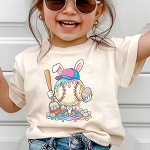 May include: A cream-colored t-shirt featuring a cartoon baseball character with bunny ears, holding a bat and a basket of eggs. The baseball is wearing a hat and shoes, with colorful drips and Easter eggs scattered around.