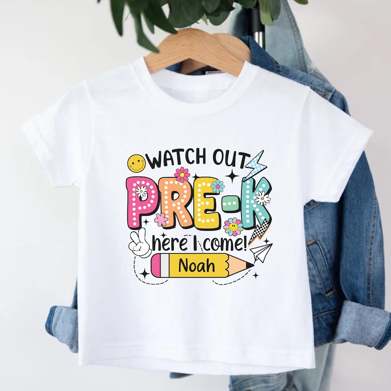 May include: White t-shirt with a colorful graphic that says "WATCH OUT PRE-K here I come!" with a pencil that says "Noah". The design includes flowers, a smiley face, and a peace sign.