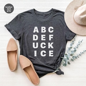 May include: Heather dark grey t-shirt with the words "ABC DEF UCK ICE" in white block letters. The shirt is styled with a pair of tan flats and a beige hat.