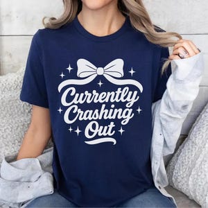 May include: Navy blue t-shirt with the text "Currently Crashing Out" in white script, featuring a bow and star accents. The shirt is a casual wear item.
