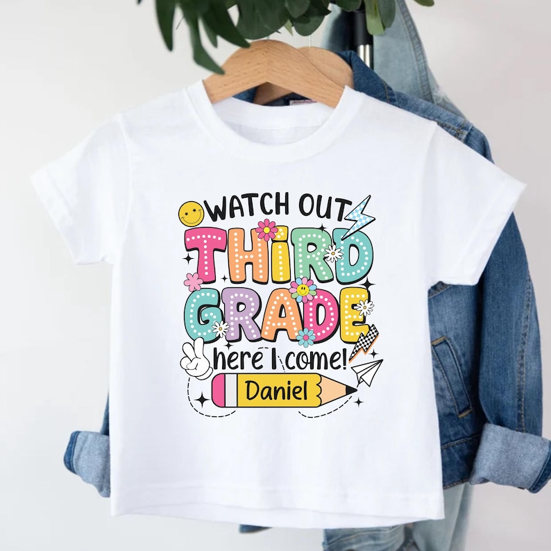 May include: White t-shirt with a colorful graphic that reads "WATCH OUT THIRD GRADE here I come!" with a pencil and the name "Daniel". The design includes flowers, a smiley face, and a peace sign.
