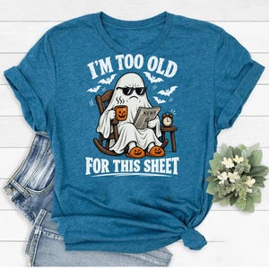 May include: A heather blue t-shirt featuring a graphic of a ghost wearing sunglasses, sitting in a rocking chair, holding a coffee mug and a newspaper, with the text I'M TOO OLD FOR THIS SHEET, surrounded by small bats and an alarm clock.