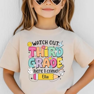 May include: A beige t-shirt with the text "WATCH OUT THIRD GRADE HERE I COME!" in colorful bubble letters. The shirt features a smiley face, flowers, a peace sign, and a pencil with the name "Ella".