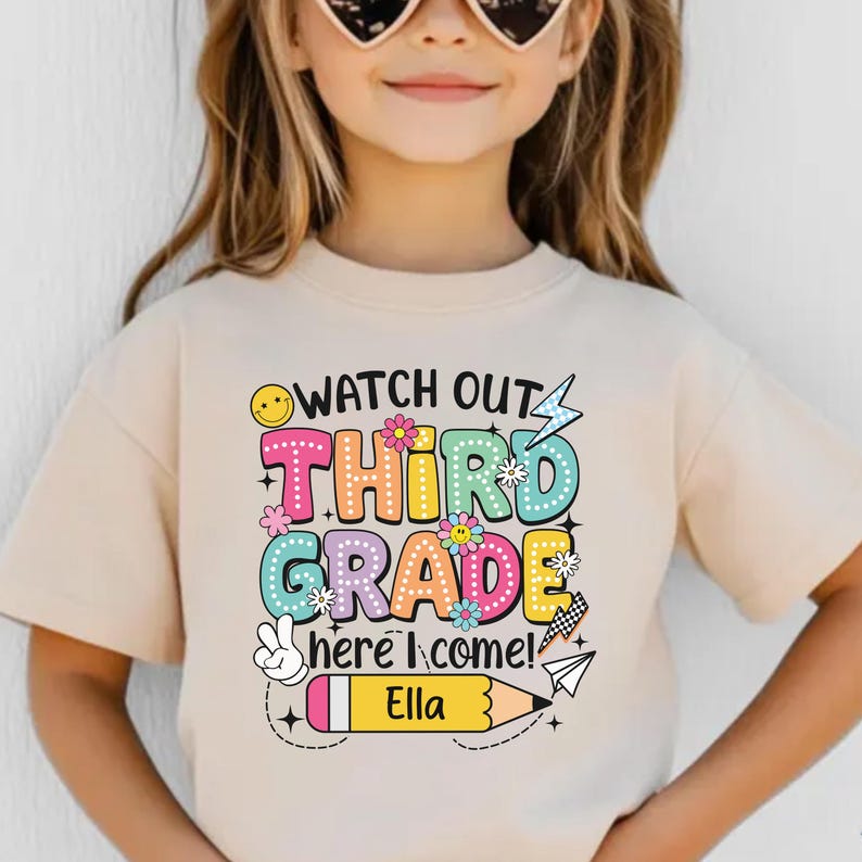 May include: A beige t-shirt with the text "WATCH OUT THIRD GRADE HERE I COME!" in colorful bubble letters. The shirt features a smiley face, flowers, a peace sign, and a pencil with the name "Ella".