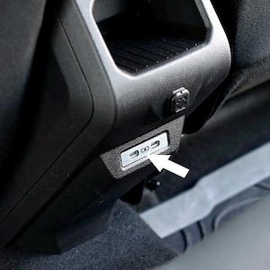 May include: Close-up of a black car interior featuring a USB charging port. The port is silver and rectangular, with two USB-C ports. The arrow points to the charging port. The interior is black.