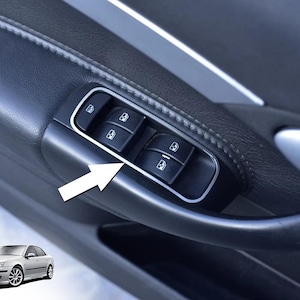 May include: Close-up of a car door panel featuring a window control cluster. The panel is black with a silver trim around the window controls. The window controls are black with white icons. The door handle is visible.