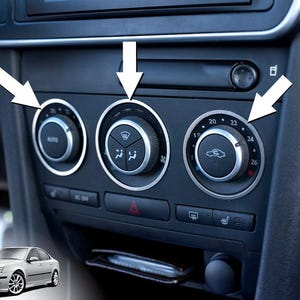 Könnte beinhalten: Nahaufnahme des Armaturenbretts eines Autos mit Klimaanlagenknöpfen. Die schwarzen Knöpfe haben silberne Zierleisten und zeigen Temperatureinstellungen und Lüftersteuerungen an. Der "AUTO"-Knopf ist sichtbar, zusammen mit dem Temperaturregler und den Lüftergeschwindigkeitsreglern.