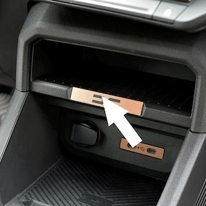 Könnte beinhalten: Nahaufnahme eines Autoinnenraums mit einem Ablagefach, das mit einem kupferfarbenen Akzent versehen ist. Auf dem Akzent ist die Schrift "E =" eingraviert. Darunter ist ein USB-C-Anschluss zu sehen.
