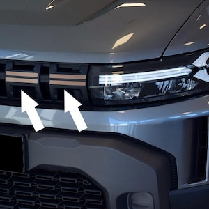 Puede incluir: Primer plano de la parrilla y el faro delanteros de un coche gris. La parrilla presenta un diseño negro con detalles en color bronce. El faro es elegante y moderno, con luces LED blancas brillantes.