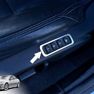 Op de afbeelding: Close-up van een autostoel met een zilverkleurig bedieningspaneel. Het paneel heeft vier zwarte knoppen met de labels 1, 2, 3 en M. De stoel is van zwart leer. Een pijl wijst naar het paneel.