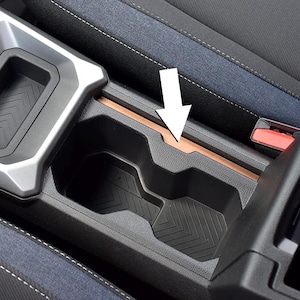 Peut inclure: Un porte-gobelet de console centrale automobile en plastique noir avec un motif à chevrons texturé sur la base et une fine bande décorative horizontale de couleur cuivre près du bord supérieur, indiquée par une flèche blanche.