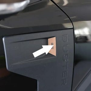 May include: A black vehicle side panel featuring the embossed word DUSTER in vertical capital letters. A matte dark gray trim piece is attached to the panel, containing a small, light brown rectangular insert marked with two horizontal lines.