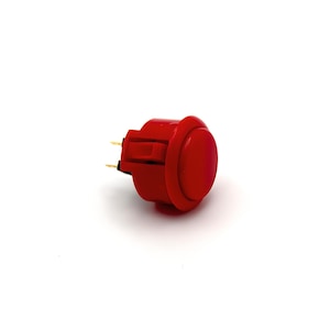 May include: A red arcade button with two gold-colored terminals. The button is round and has a smooth surface.