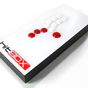 Puede incluir: Un stick de arcade blanco con botones rojos y blancos. El stick tiene una base negra y el texto "Hit Box" impreso en el lateral.