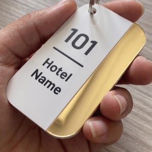 Può includere: Un portachiavi rettangolare per hotel con una sezione superiore bianca e una sezione inferiore dorata. L'etichetta ha un portachiavi argentato attaccato. Il testo "101" e "Hotel Name" è stampato in nero sulla sezione bianca.