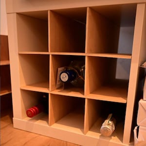Puede incluir: Una estantería de madera blanca con nueve compartimentos, algunos de los cuales contienen botellas de alcohol. La estantería está hecha de madera y tiene un acabado de madera clara.