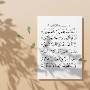 May include: A white rectangular print featuring Arabic calligraphy. The text is in black and is arranged in multiple lines. The print is set against a beige background with shadows and green leaves.