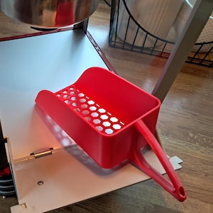 May include: A red popcorn scoop with a handle and a perforated bottom sits on a white surface. The scoop is part of a popcorn machine, with a stainless steel pot and a wooden door visible.