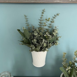 May include: A white, textured wall planter filled with green and gray faux eucalyptus and small white flowers. The planter is mounted on a light blue wall. Decorative home decor.