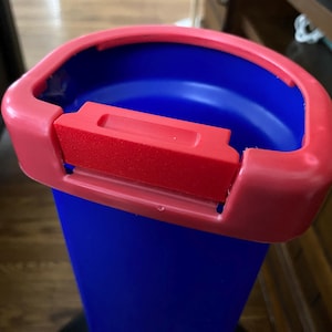 May include: A blue plastic bin with a red lid and a red handle. The bin has a smooth surface and is designed for recycling.