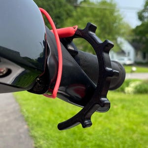 May include: A black plastic throttle assist device attached to a motorcycle handlebar grip. The accessory features a gear-like circular design with a protruding lever arm and is secured in place by a thin, bright red elastic silicone band.