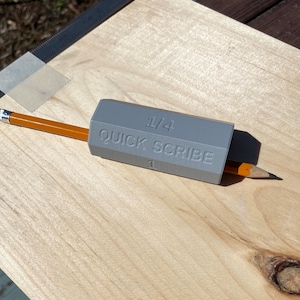 May include: A gray 3D printed tool labeled "1/4 QUICK SCRIBE" with a pencil inserted into it. The tool is lying on a light wood surface.