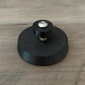 May include: A black, circular object with a raised, cylindrical top. A silver screw is visible on the top. The object appears to be made of a textured, matte material. The base is approximately 3.8 cm in diameter.