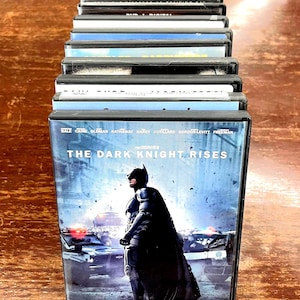 May include: A vertical stack of DVD cases arranged in a single row. The front-most case features the movie title The Dark Knight Rises, displaying a graphic of Batman standing before police vehicles, with the text A monumental conclusion to the epic trilogy.