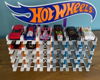 May include: A white and gray Hot Wheels car display stand with six toy cars. The stand has a blue and orange Hot Wheels logo. The cars are red, blue, pink, black, black, and white.