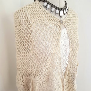 May include: A cream-colored crocheted shawl with a delicate openwork pattern. The shawl has a button closure at the neck and a scalloped edge.