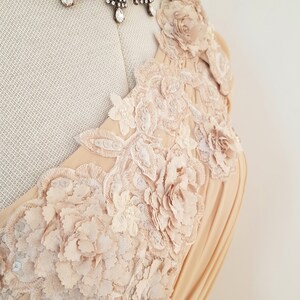 May include: A beige dress with a one-shoulder neckline and intricate floral lace detailing. The lace features delicate flowers and leaves, creating a romantic and elegant look.