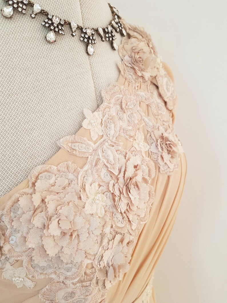 May include: A beige dress with a one-shoulder neckline and intricate floral lace detailing. The lace features delicate flowers and leaves, creating a romantic and elegant look.
