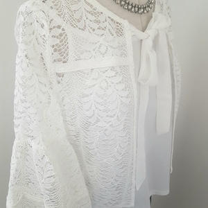 以下が含まれることがあります： ボウタイディテールが付いた白いレースのショール。ショールは、波状の縁取りが施され、繊細なレースで作られています。