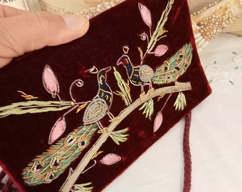 May include: A small, red velvet bag with a drawstring closure. The bag is decorated with embroidered peacocks and floral designs in gold, green, and pink thread.