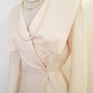 May include: A white, long-sleeved blouse with a large, pointed collar and a single button fastening. The blouse has a fitted waist and a slightly flared hem. The blouse is worn with a pearl necklace.