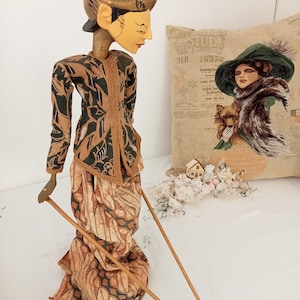 May include: A traditional Indonesian puppet with a carved wooden head and painted face. The puppet is wearing a green and brown patterned jacket and a long, flowing skirt with a batik design. The puppet is holding two wooden sticks.