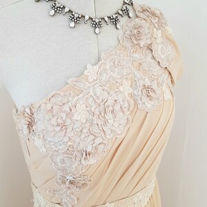 May include: A one-shoulder, floor-length dress in a pale beige colour. The dress features delicate floral lace appliques and a beaded belt.