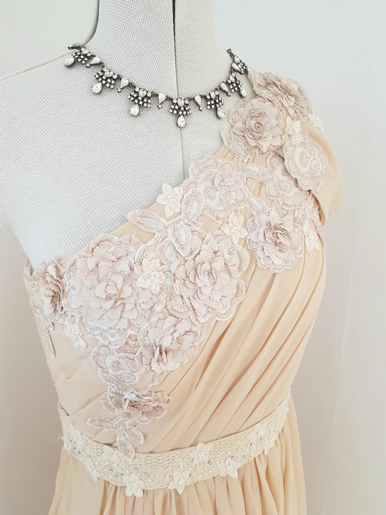 May include: A one-shoulder, floor-length dress in a pale beige colour. The dress features delicate floral lace appliques and a beaded belt.