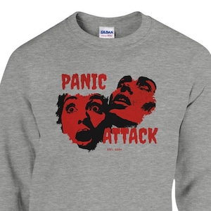 Könnte beinhalten: Ein graues Sweatshirt mit Rundhalsausschnitt und einem roten Aufdruck von zwei Gesichtern mit offenem Mund und den Worten "PANIC ATTACK" in Rot. Der Text "EST. 2020" befindet sich unter der Grafik. Der Markenname "GILDAN" ist am Kragen sichtbar.