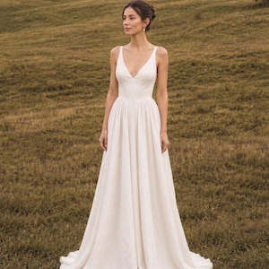Puede incluir: Un vestido de novia blanco con escote en V y falda fluida en línea A. El vestido tiene un corpiño ajustado y una falda amplia que llega hasta el suelo. El vestido está hecho de una tela suave y texturizada. La modelo está en un campo.