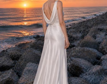 May include: A floor-length white satin gown featuring a sleeveless bodice with a deep cowl back neckline, a fitted waist, and a flowing skirt that pools slightly at the hem, crafted from a smooth, lustrous fabric with a subtle sheen.