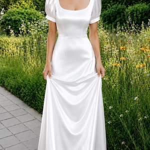 May include: A floor-length white satin dress featuring short puff sleeves, a square neckline, and a fitted bodice that transitions into a flowing, floor-length skirt. The fabric has a smooth, lustrous finish that catches the light.