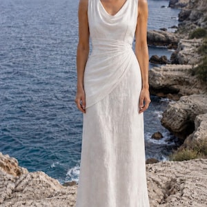 K&ouml;nnte beinhalten: Ein &auml;rmelloses, bodenlanges wei&szlig;es Kleid mit einem drapierten Wasserfallausschnitt. Das Kleid hat ein figurbetontes Oberteil mit einem gewickelten Detail an der Taille und einen flie&szlig;enden Rock. Das Model steht auf einer felsigen Klippe mit Blick auf das Meer.