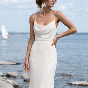 May include: A white, floor-length dress with a cowl neckline and spaghetti straps. The dress is made of a lightweight fabric and has a fitted silhouette. The model is standing near a body of water with a sailboat in the background.