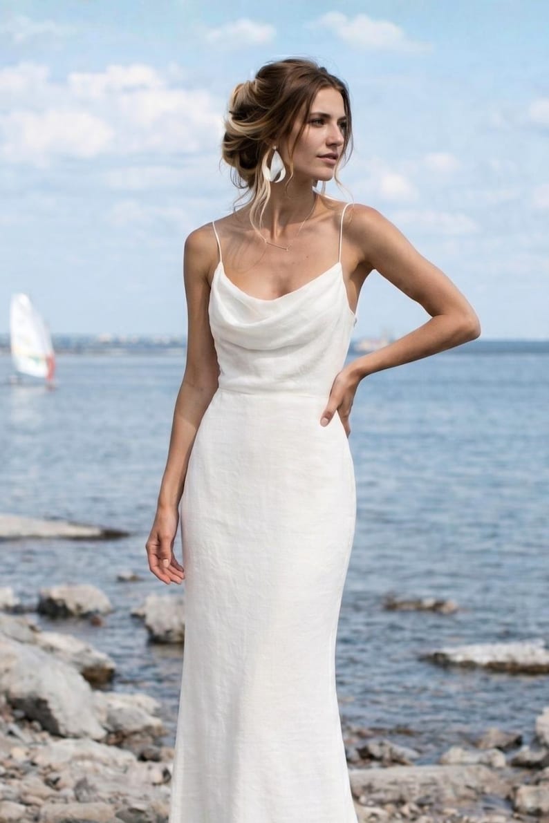 May include: A white, floor-length dress with a cowl neckline and spaghetti straps. The dress is made of a lightweight fabric and has a fitted silhouette. The model is standing near a body of water with a sailboat in the background.