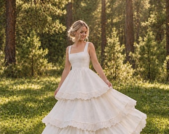 May include: A floor-length, off-white tiered dress featuring a square neckline, thick shoulder straps, and a fitted bodice. The skirt consists of three distinct layers, each finished with a wide, intricate lace trim along the bottom hem.